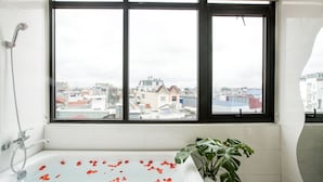 A bathroom with a bathtub, a shower, and a large window with a view of buildings. - Aui Hotel (Hai Phong)