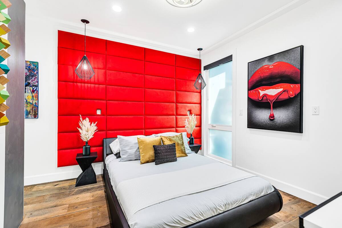 A modern bedroom with a red padded headboard, a large bed with multiple pillows, a black side table with a vase, and a painting of lips on the wall.