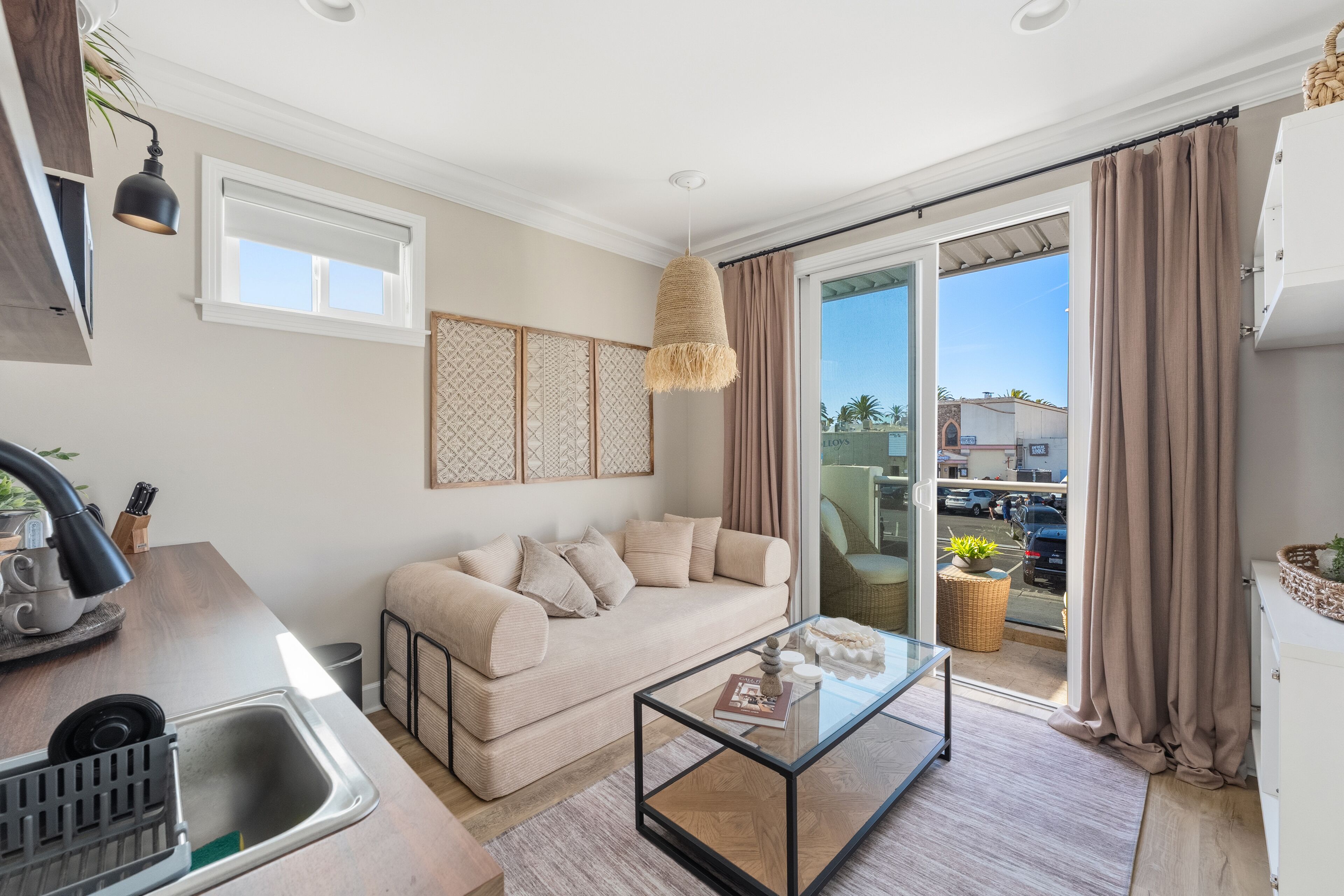 A modern living room with a sofa, a glass coffee table, a kitchen area with a sink, and a sliding glass door leading to an outdoor area.