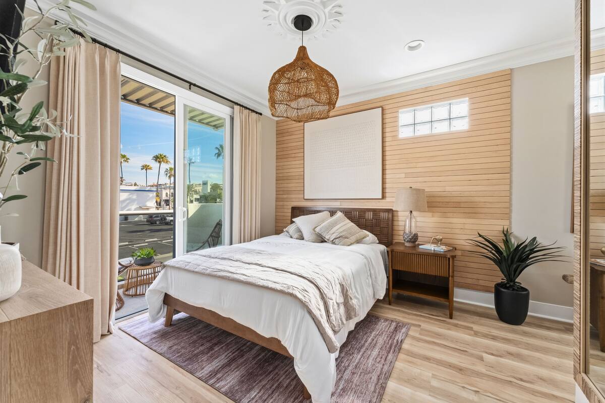 A bedroom with a bed, bedside table, lamp, potted plant, and a view of the outside.
