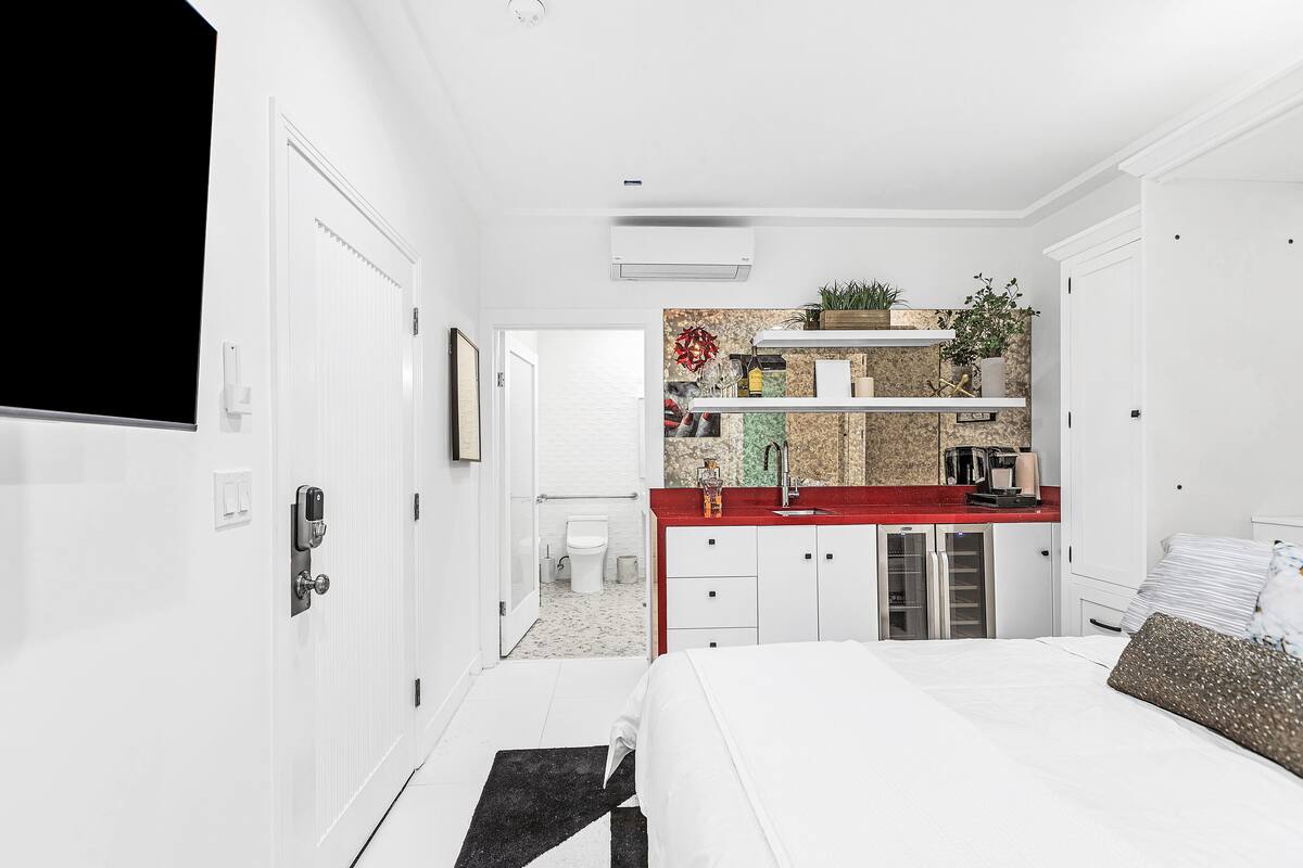 A modern hotel room with a white bed, a red accent wall, a kitchen area with a sink and countertop, and a small bathroom visible through an open door.
