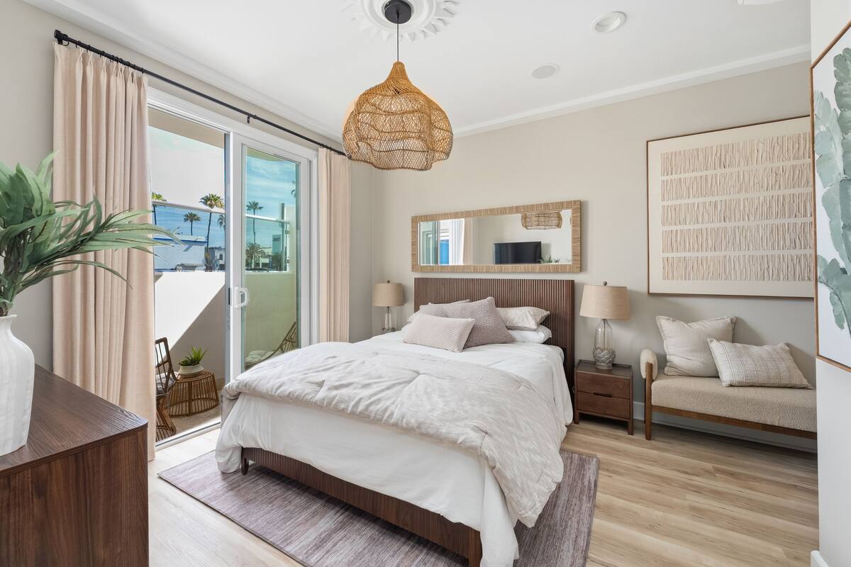 A bedroom with a bed, bedside tables, a hanging light fixture, a large mirror, and a view through the glass door.
