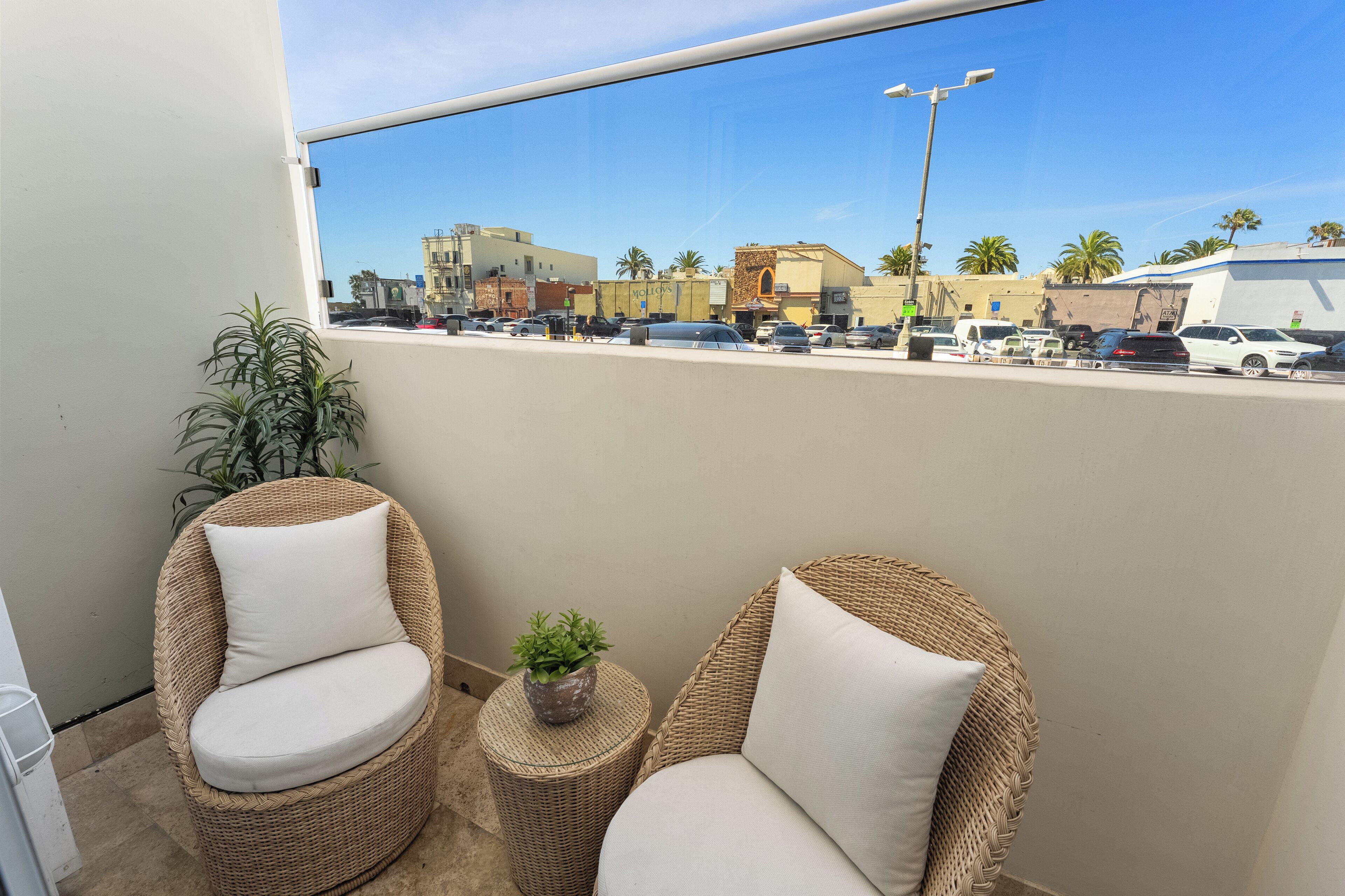 A balcony with wicker chairs and a small round table with a plant, overlooking a parking lot and buildings.