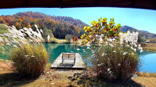 Une vue sur le lac, avec un quai et une chaise, le tout entouré d’un feuillage automnal et d’une colline boisée.