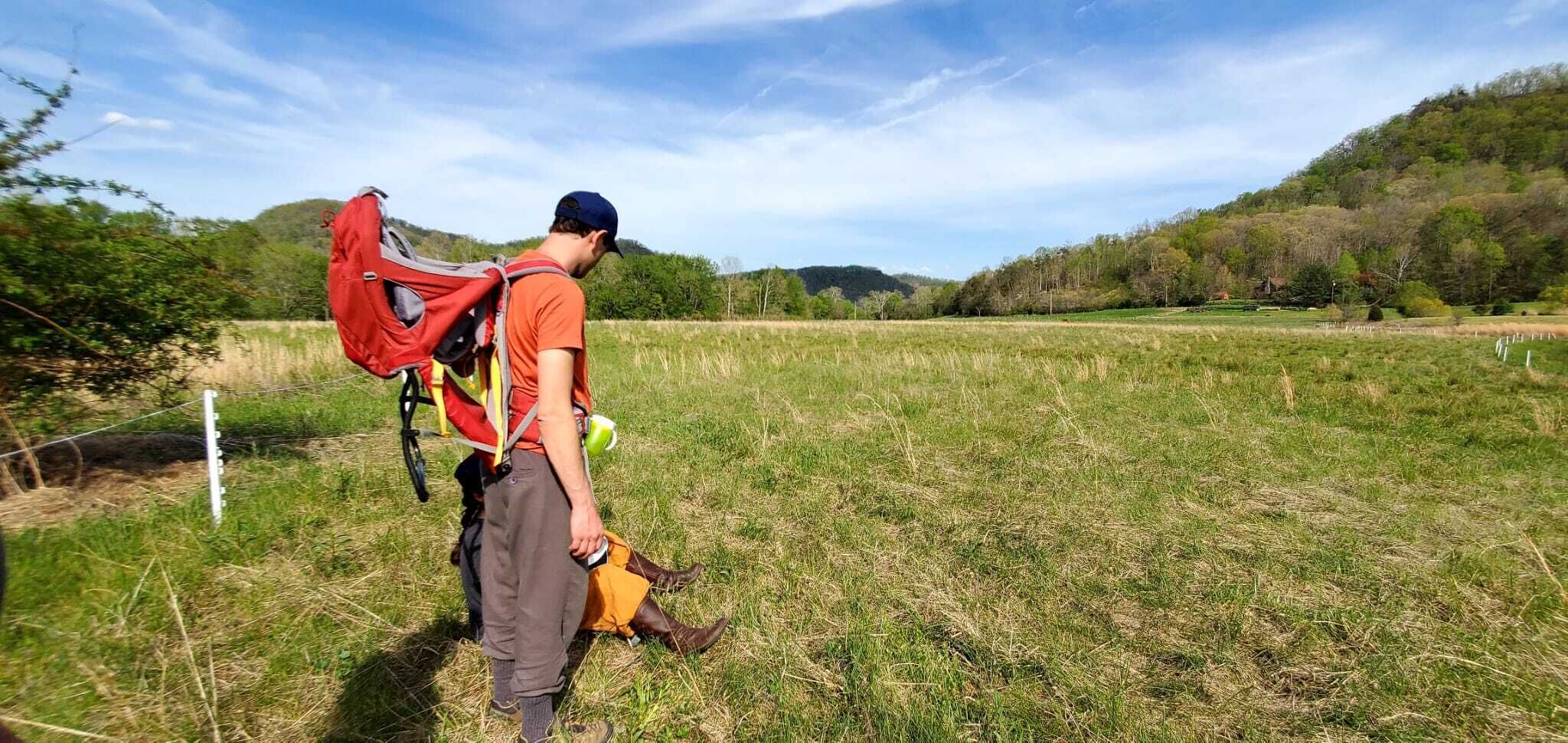Une personne se tient dans un champ, portant un sac à dos rouge et un sac jaune, près d’une clôture.