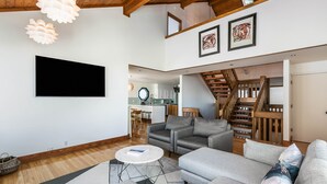 Ein modernes Wohnzimmer mit einem Flachbildfernseher, grauen Sofas, einem runden Couchtisch und einer Treppe, die in eine obere Etage führt.