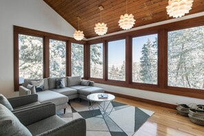 A modern living room with a sectional sofa, a coffee table, and large windows offering a view of snow-covered trees.