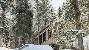 Eine Holzhütte in einem verschneiten Wald mit roter und weißer Fassade, umgeben von hohen Nadelbäumen.