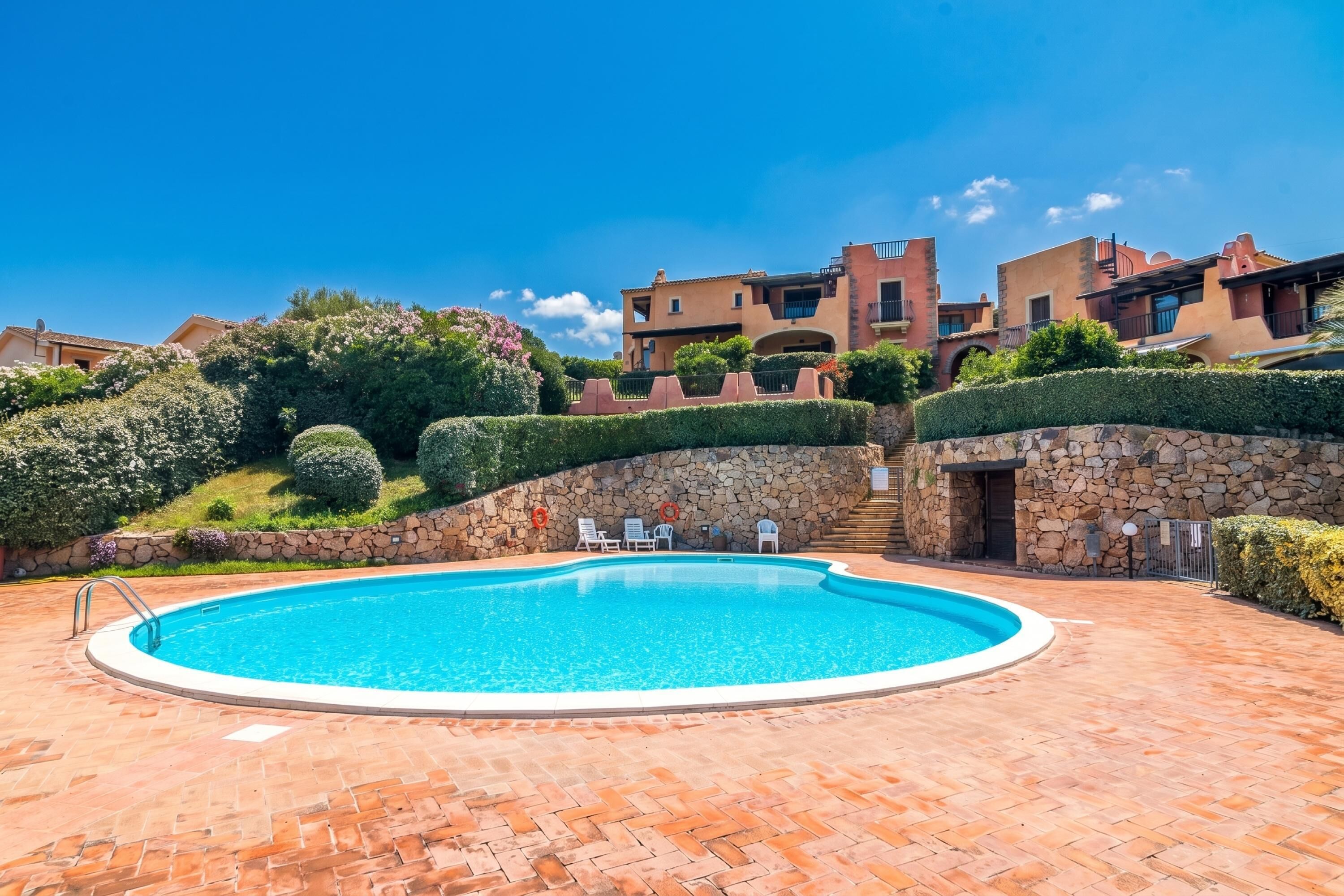 A swimming pool with a stone wall, a stone staircase, and a view of buildings in the background.