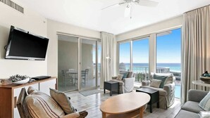 Ett rymligt vardagsrum med en platt-TV, ett soffbord i trä och en soffa. Det finns en balkong med utsikt över havet.