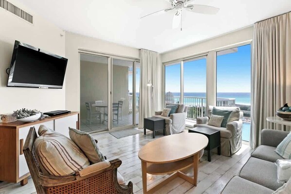 A spacious living area with a flat-screen TV, a wooden coffee table, and a sofa. There is a balcony with a view of the ocean.