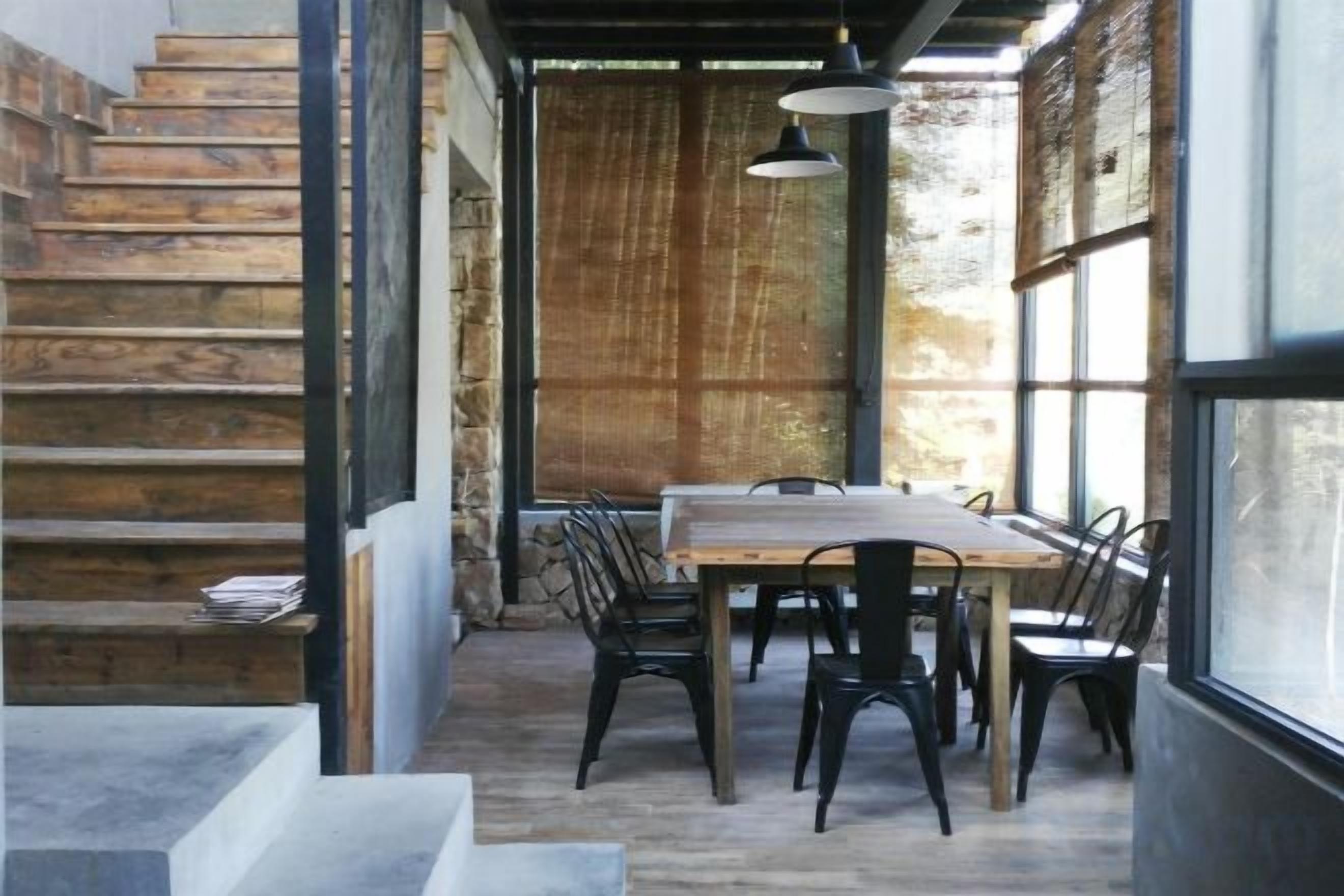 Un comedor con mesa y sillas de madera, una escalera y ventanas grandes.