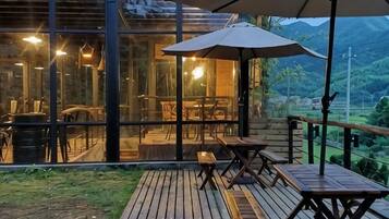 Un área de comedor al aire libre con mesas y sillas, una terraza de madera y un muro de vidrio con vista a un paisaje montañoso.