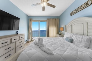 A hotel room with a bed, a ceiling fan, a TV, a dresser, a nightstand, and a view of the ocean.