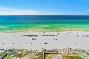 An aerial view of a beach with clear turquoise waters, a sandy shore, and several beach umbrellas and lounge chairs.