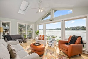 Relax in this spacious living area with a high ceiling and large windows offering a view of a body of water and a distant shoreline. The room features a sofa, armchair, and a rug, perfect for unwinding.