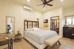 A bedroom with a bed, a ceiling fan, an air conditioning unit, a bedside table with a lamp, a bench, a dresser, and a mirror.