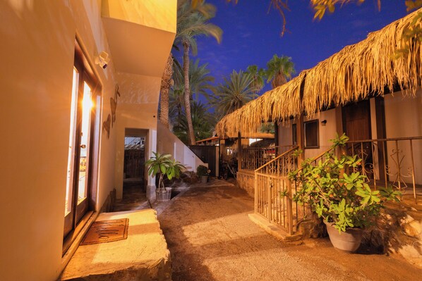 A narrow pathway leading to a building with a thatched roof, surrounded by palm trees and potted plants.
