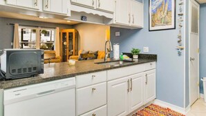 A kitchen with white cabinets, a microwave, and a sink. There is a window with curtains, a painting on the wall, and a colorful rug. - Beachside Townhouse w/ Community Pool & Patio (Pensacola)