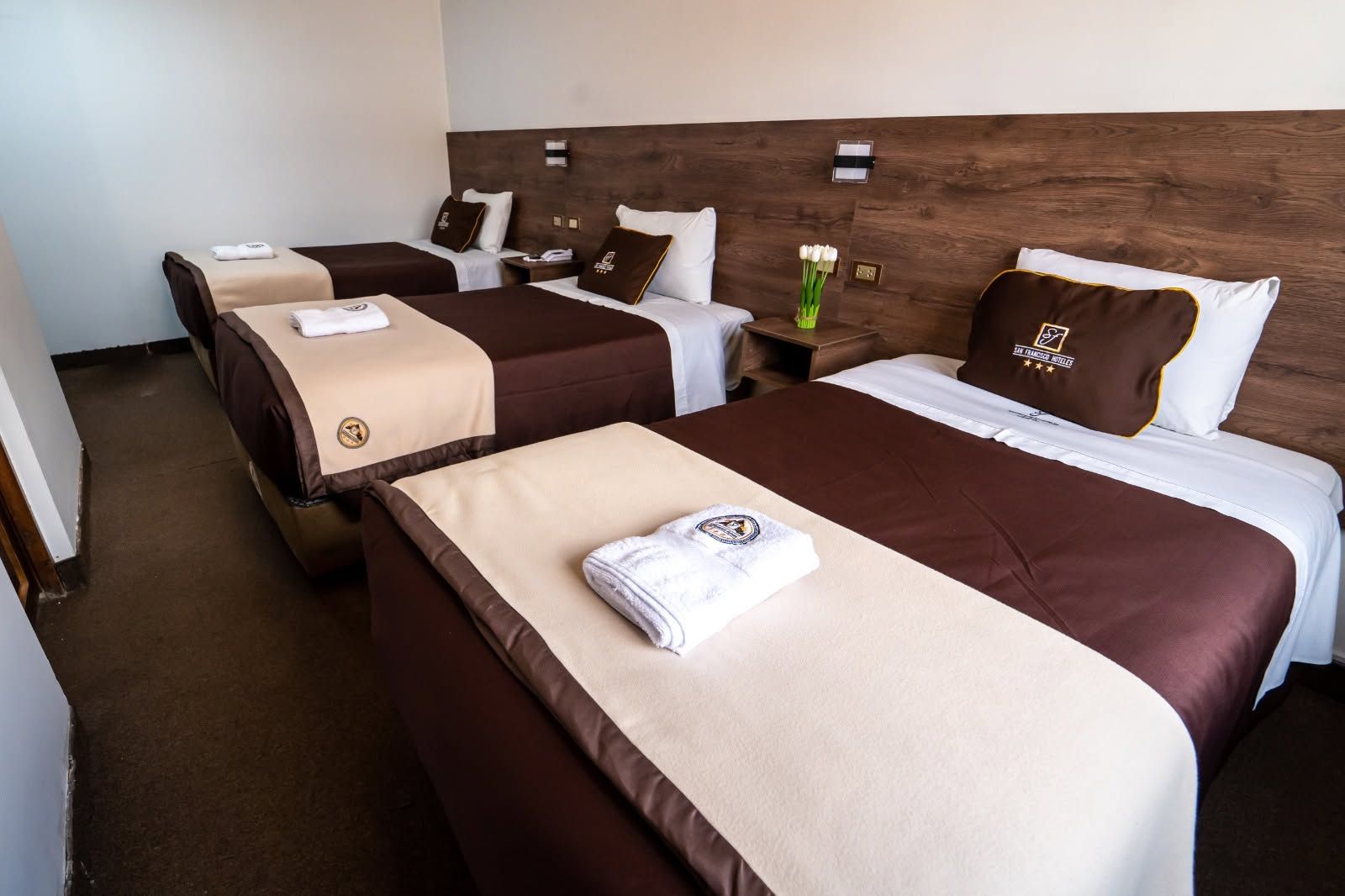 A hotel room with three beds, each with a brown headboard and white bedding. There are towels on the beds, and a vase with flowers on the nightstand.