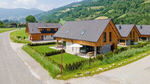 A row of modern wooden houses with sloped roofs, a paved road, and a green landscape with hills in the background.