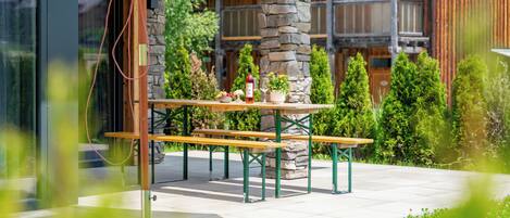 An outdoor dining area with a wooden table and benches, surrounded by greenery and a stone pillar.