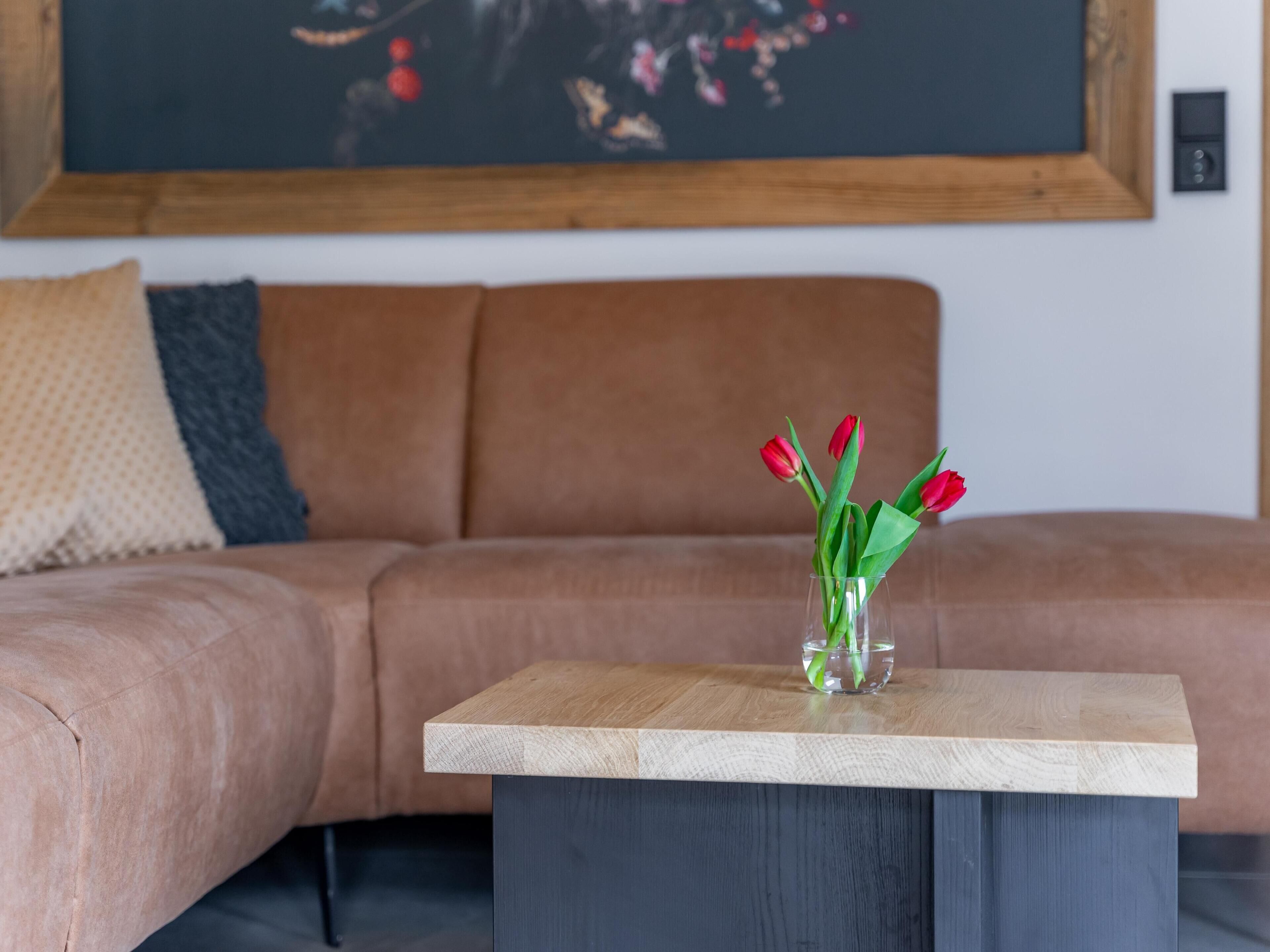 A living room with a beige sofa, a glass vase with red flowers on a wooden coffee table, and a painting on the wall.