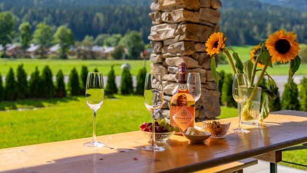 A table with wine glasses, a bottle of Prosecco Rosé, grapes, and a bowl of nuts, set against a scenic mountain backdrop.