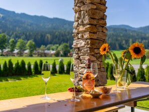 A table with wine glasses, a bottle of Prosecco Rosé, grapes, and a bowl of nuts, set against a scenic mountain backdrop.