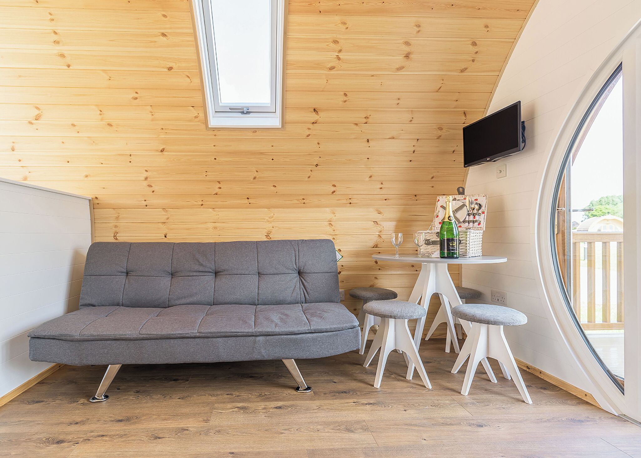 Une pièce chaleureuse avec un canapé gris, une petite table sur laquelle se trouvent une bouteille et des verres, et un téléviseur fixé au mur.