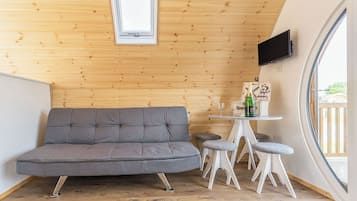 Une pièce chaleureuse avec un canapé gris, une petite table sur laquelle se trouvent une bouteille et des verres, et un téléviseur fixé au mur.
