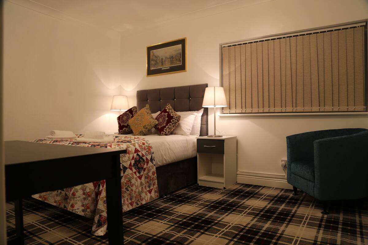 A hotel room with a bed, bedside tables, a desk, a chair, a window with blinds, and a framed picture on the wall.