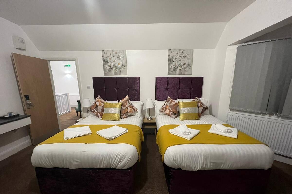 A hotel room with two beds, each with a purple headboard and white bedding. There are two bedside tables with lamps, and a window with curtains.