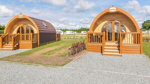 Two wooden arched cabins with wooden railings and steps, set on a gravel path with grass in between.