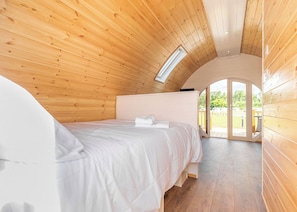 Une chambre avec un plafond en bois, une lucarne, un lit avec une literie blanche et une vue sur l’extérieur par une porte ouverte.