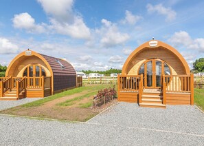 Deux cabanes en bois à toit en arc, dotées de balustrades et d’escaliers en bois, sont construites le long d’un chemin de gravier, avec de l’herbe qui pousse entre les pierres.