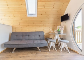 Une pièce chaleureuse avec un canapé gris, une petite table ronde sur laquelle se trouvent une bouteille et des verres, un téléviseur fixé au mur et une grande fenêtre.