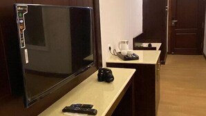 A hotel room with a large flat-screen TV on a wooden stand, a small table with a camera and remote controls, and a door leading to another room. - Hotel Barath Ladakh (Leh)
