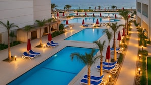 A pool area with multiple pools, lounge chairs, and umbrellas, surrounded by palm trees and a view of the ocean. - Porto Said Beach Escape (Al-Manasrah)