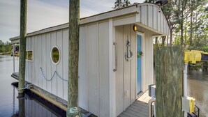 Ein kleines, weißes Hausboot an einem Steg mit einer blauen Tür, einem kleinen Fenster und einem Deck.