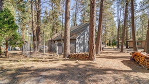 A rustic cabin surrounded by tall pine trees, with a pile of firewood and a fenced area. - Graeagle Vacation Rental Cabin w/ Game Room! (Graeagle)