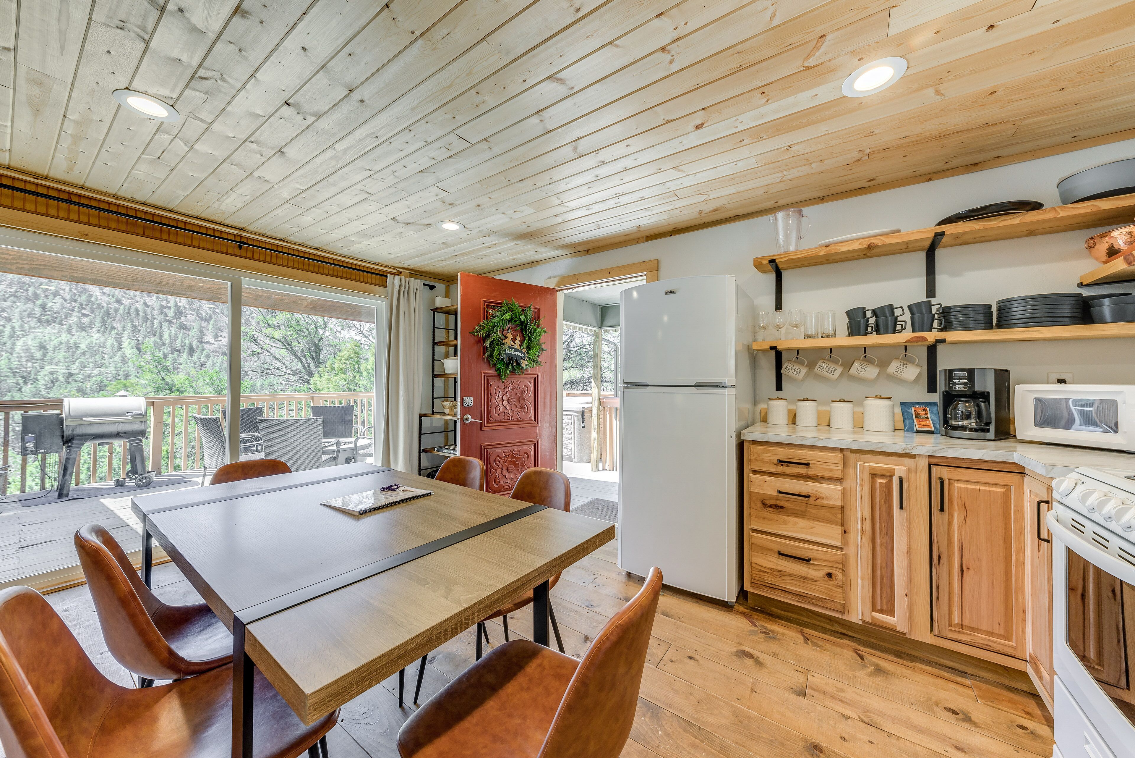A wooden deck with a table and chairs, a kitchen with wooden cabinets and appliances, and a large window overlooking greenery.