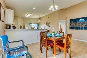 Uma sala de jantar com mesa e cadeiras de madeira, cozinha ao fundo e um quadro na parede.