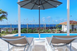 Zona junto a la piscina con sillones de descanso y una sombrilla, con vista a una piscina azul cristalina y al océano.