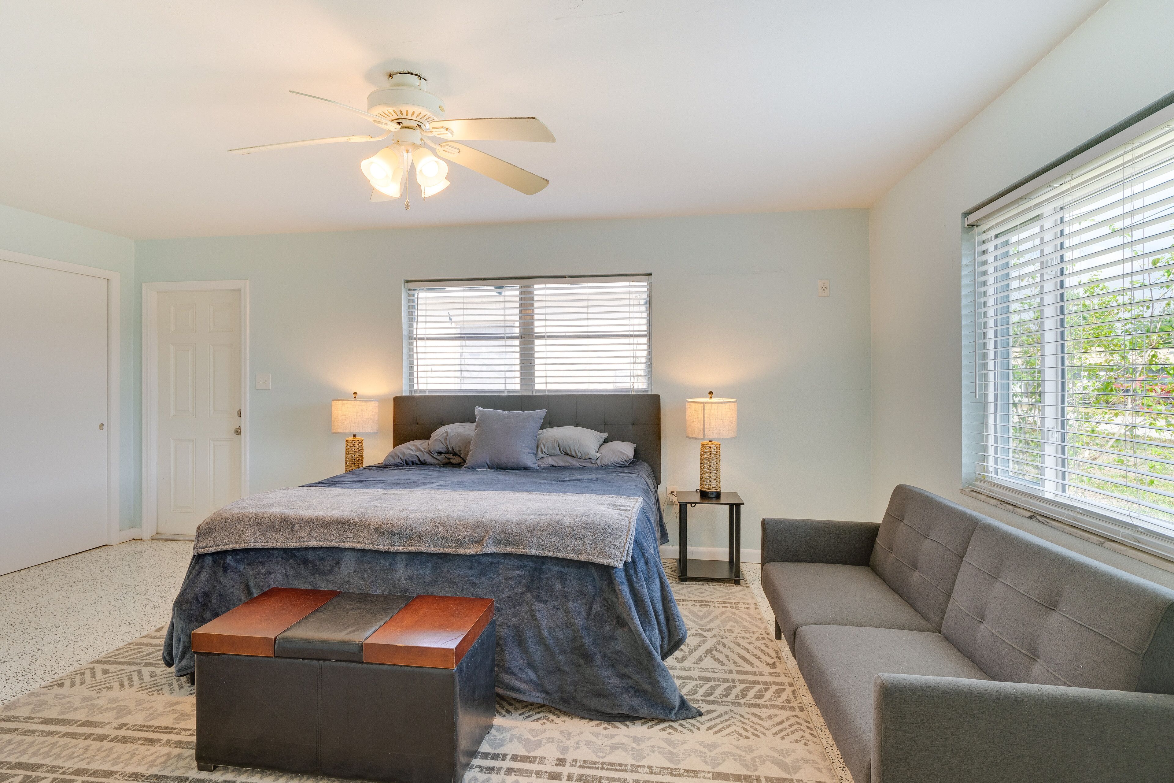 Une chambre à coucher comprenant un lit, des lampes de chevet, un ventilateur de plafond, une fenêtre avec des stores, un canapé et un tapis à motifs.