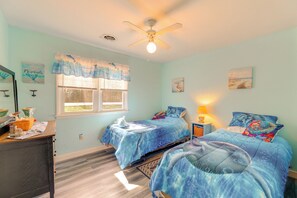 Une chambre avec deux lits, un ventilateur de plafond, une fenêtre avec des rideaux, un bureau avec une lampe et des œuvres d’art accrochées aux murs.