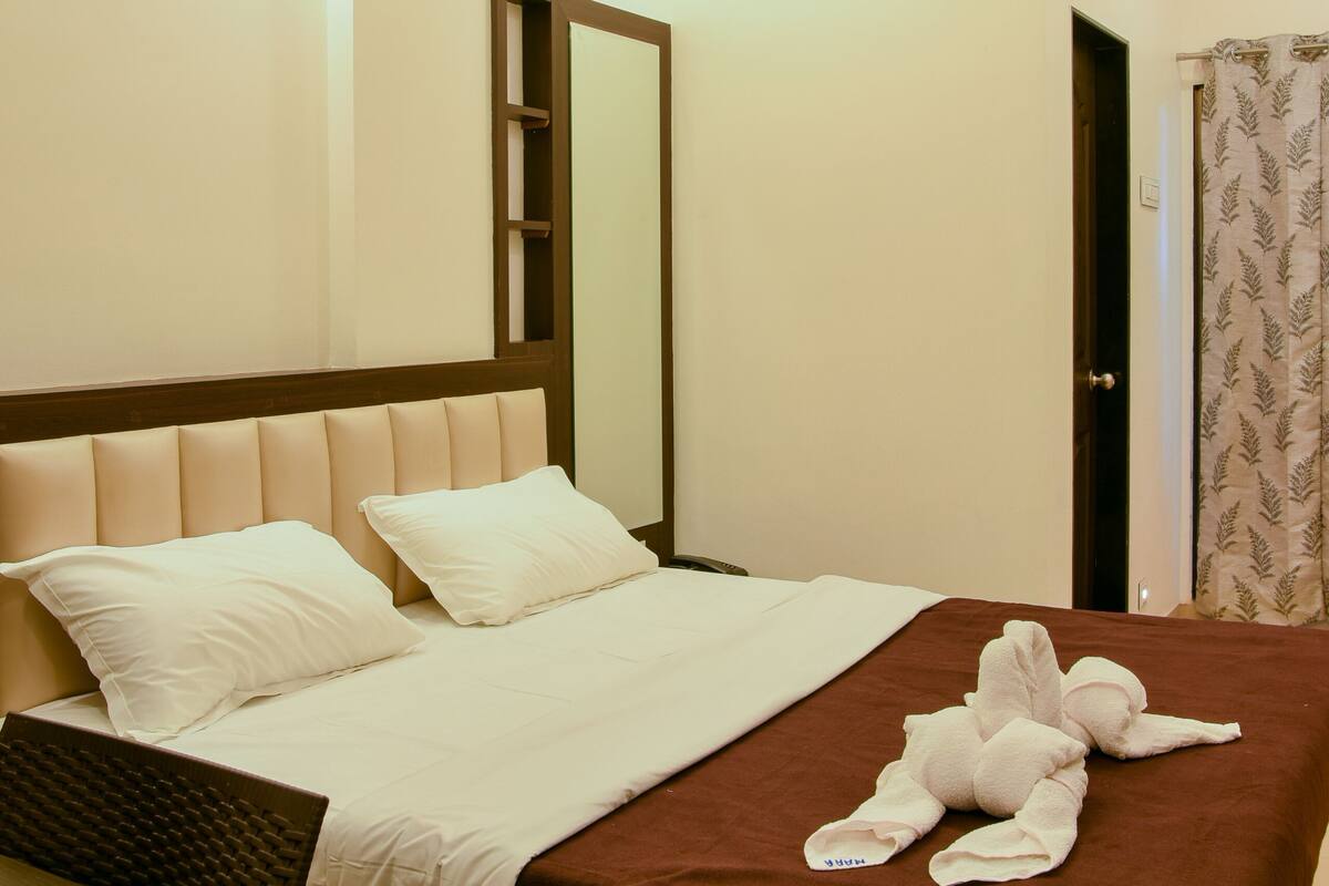 Cama bem arrumada com colcha marrom e almofadas brancas, cabeceira de madeira e mesa de cabeceira com abajur.