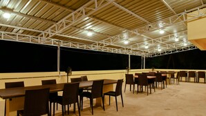 An outdoor covered dining area with multiple tables and chairs, a high ceiling with metal beams, and a dark background suggesting nighttime. - Malvan's Aishwarya Hotel (Malvan)