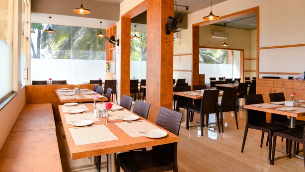 A restaurant with multiple dining tables and chairs, large windows with a view of palm trees, and pendant lighting. - Malvan's Aishwarya Hotel (Malvan)