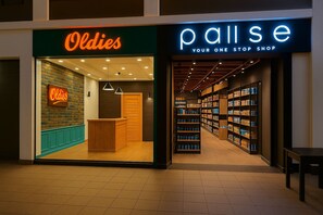 A store named 'Oldies' with a wooden counter and shelves filled with products. Adjacent to it is 'Pallise Your One Stop Shop' with a variety of items on shelves.